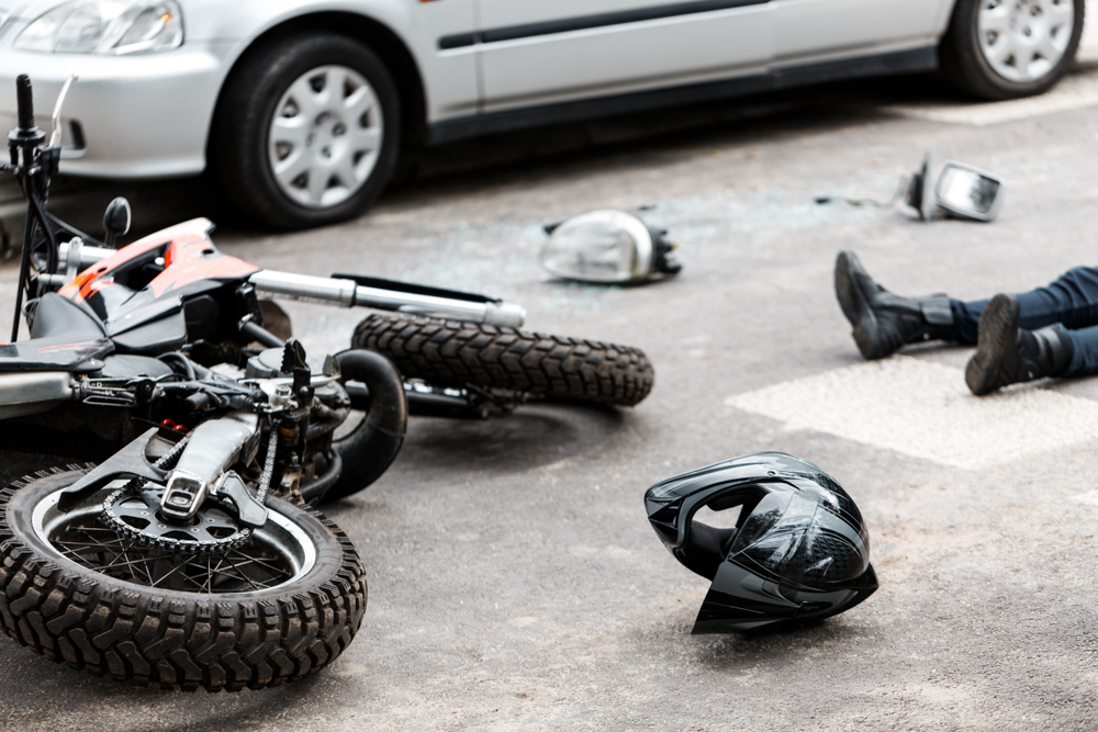 Motorcycle crash scene on city street with fallen bike, helmet on the ground, and injured rider near a car after traffic accident.