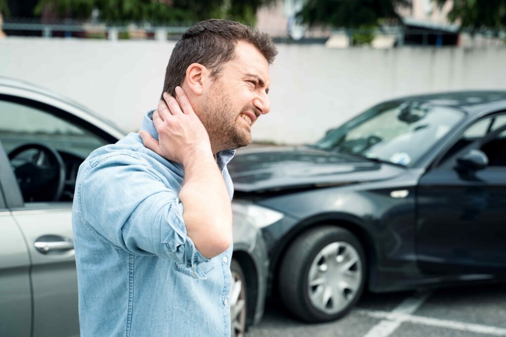 Injured driver holding neck in pain after car accident, standing near damaged vehicles on city street.