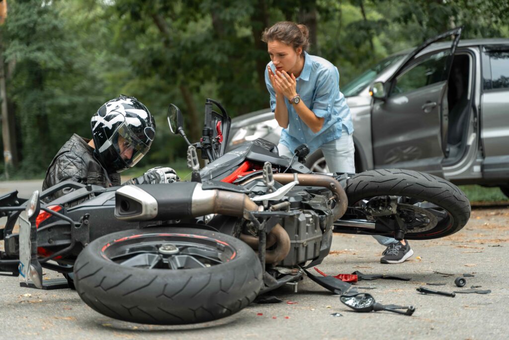 Motorcycle accident scene with injured rider on the ground and concerned woman calling for help beside a damaged bike and car on a wooded road. Motorcycle accident scene with injured rider on the ground and concerned woman calling for help beside a damaged bike and car on a wooded road.