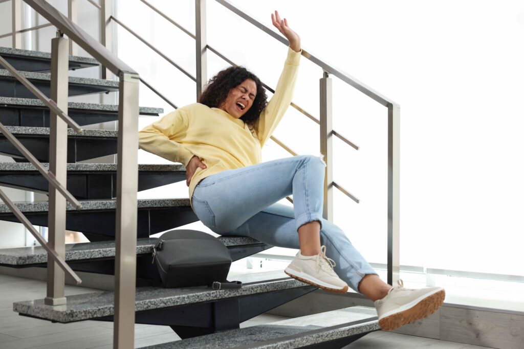 Woman grimacing in pain after slipping and falling on indoor staircase, holding her lower back in a home accident injury scene.