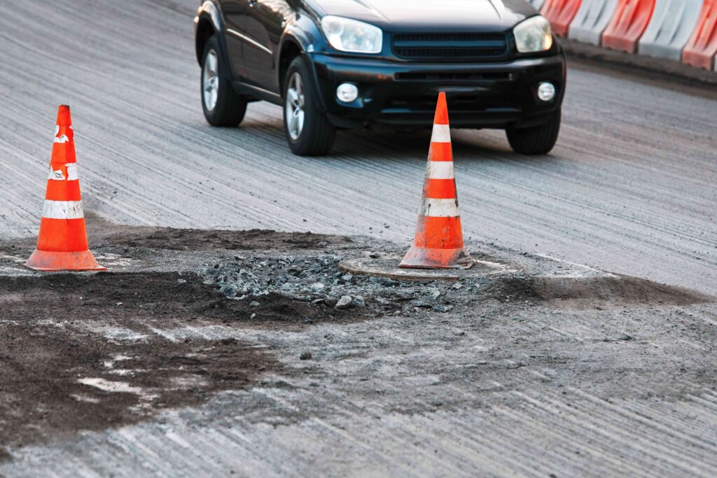 Pothole on damaged road marked with traffic cones as car approaches, highlighting road hazard and safety risk