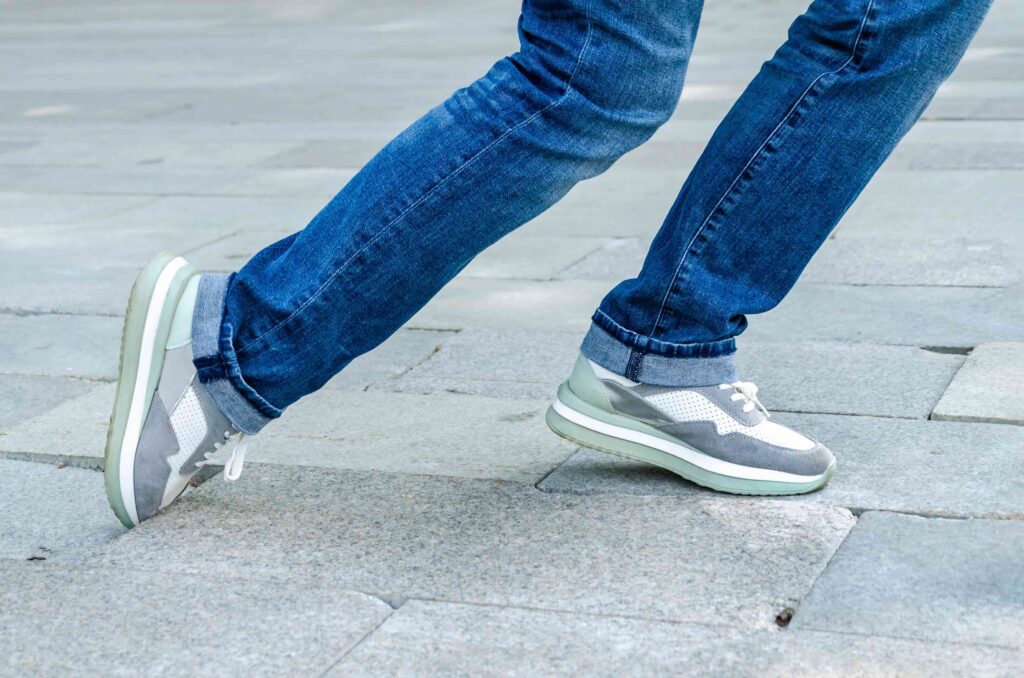 Person walking on pavement wearing blue jeans and casual sneakers, close-up of legs in motion Person walking on pavement wearing blue jeans and casual sneakers, close-up of legs in motion