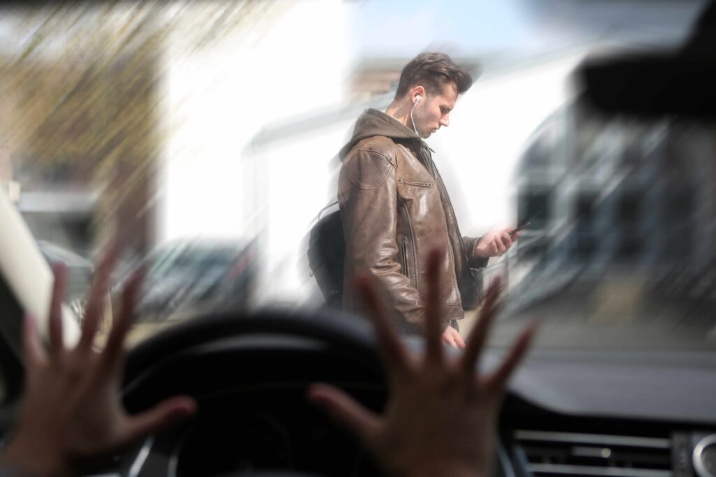 Distracted pedestrian using smartphone while crossing street as driver reacts inside car to avoid accident